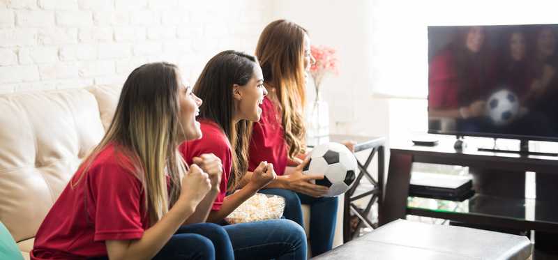 Teen girls watching soccer on tv while holding a soccer ball.