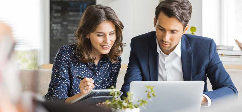 Two people in office looking at laptop while one also takes notes.