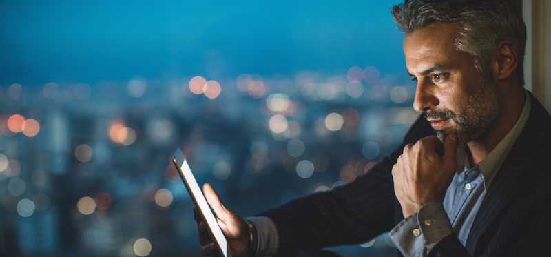 Businessperson looking at tablet against backdrop of brightly lit city after dark.