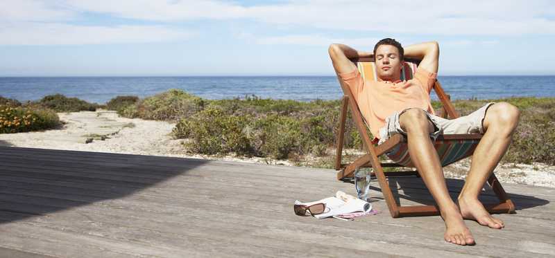Person sitting on a deck chair and napping in the sun, with the beach and ocean in the background.