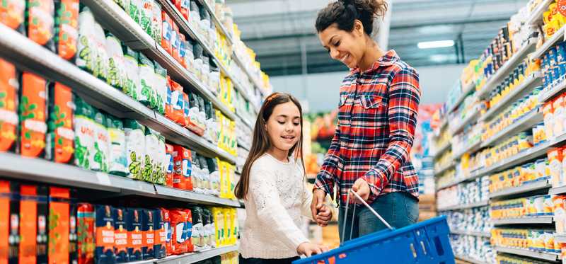 Parent and child browsing grocery aisle and placing items in blue shopping basket.