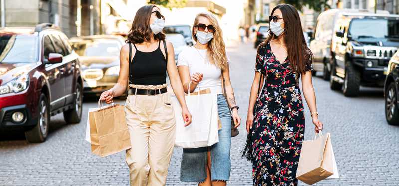 Three women wearing masks carry shopping bags down cobblestoned street.
