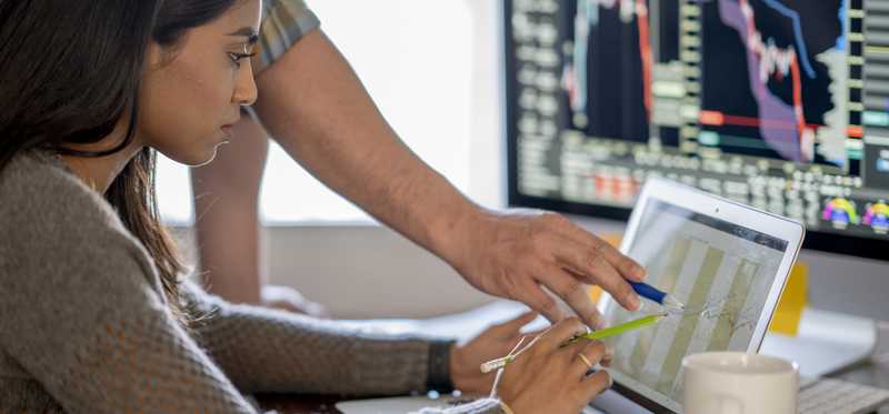 Person sitting at desk with monitors showing stock charts is conferring with another person.