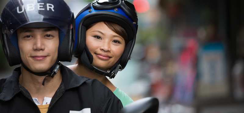 Woman rides on back of motorbike behind man with Uber helmet.