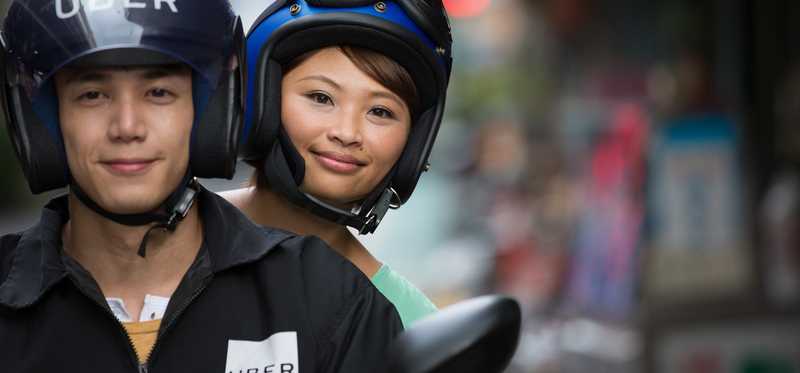 Passenger rides on back of motorbike behind driver with Uber helmet.