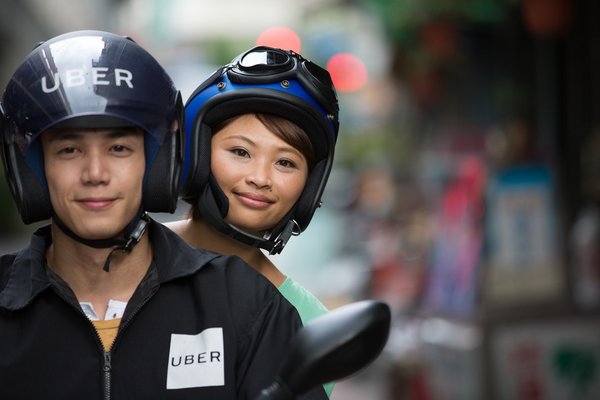 Passenger rides on back of motorbike behind driver with Uber helmet.
