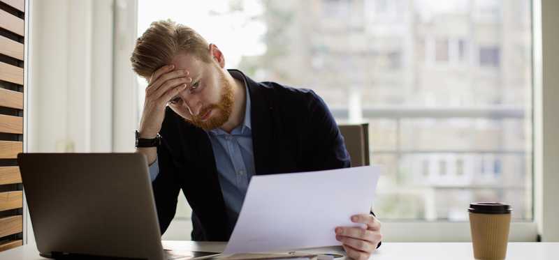 Person holds forehead in distress while looking at paperwork at desk.