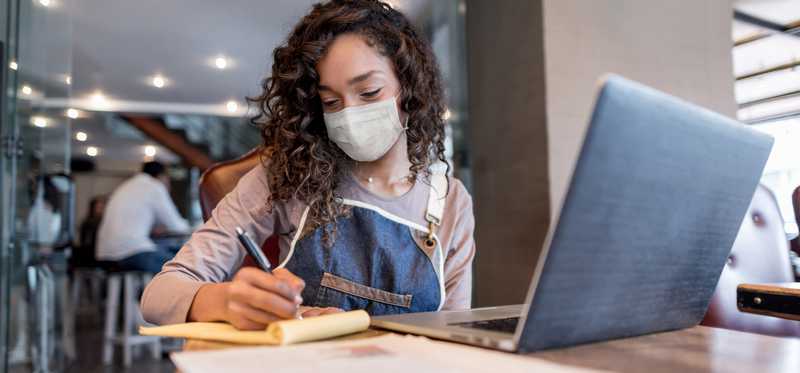 Person in face mask takes notes next to open laptop.