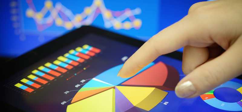 Person reviewing pie chart data on tablet against backdrop of moving stock prices.