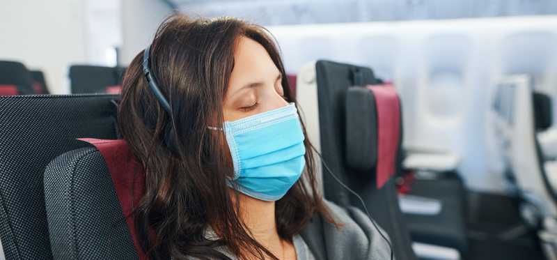 Woman wearing blue mask sitting in airplane seat with her eyes closed