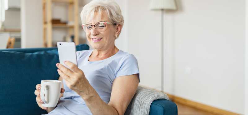 Smiling person holding coffee mug and looking at phone while sitting on couch.