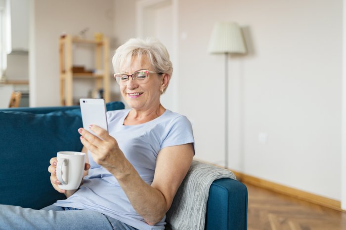 Smiling person holding coffee mug and looking at phone while sitting on couch.