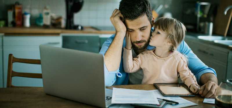 Stressed person looking at laptop and documents with young child sitting on lap kissing their face.