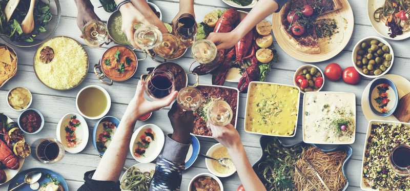 Arms reach across a dinner table full of food to toast with glasses.