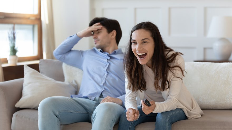 Two people sharing a couch and displaying opposite emotions. One is excited, and the other is not.