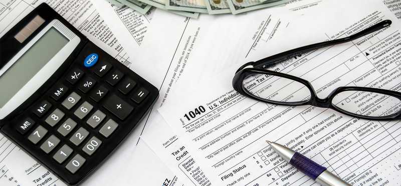 Tax forms laying scattered with a calculator, glasses, some cash, and a pen on top.