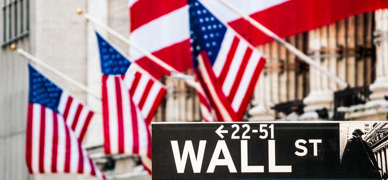 The facade of the New York Stock Exchange draped in a large American flag, and the Wall St. street sign in the foreground.