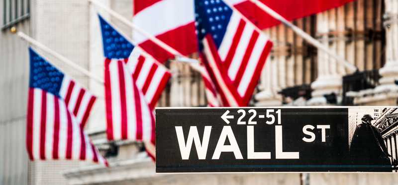 The facade of the New York Stock Exchange draped in American flags, and the Wall St. street sign in the foreground.
