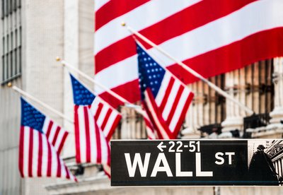 The facade of the New York Stock Exchange draped in a large American flag, and the Wall St. street sign in the foreground.