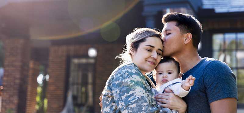 A military couple is cuddling with their baby in front of a home.