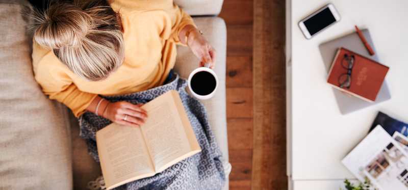 Woman holding coffee mug and reading book on couch
