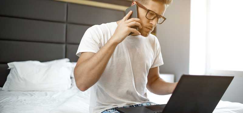 A man uses a laptop and a phone while on a bed in pajamas.