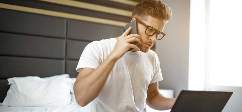 A person uses a laptop and a phone while on a bed in pajamas.