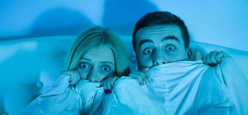 Couple in bed holding blankets to their faces, scared.