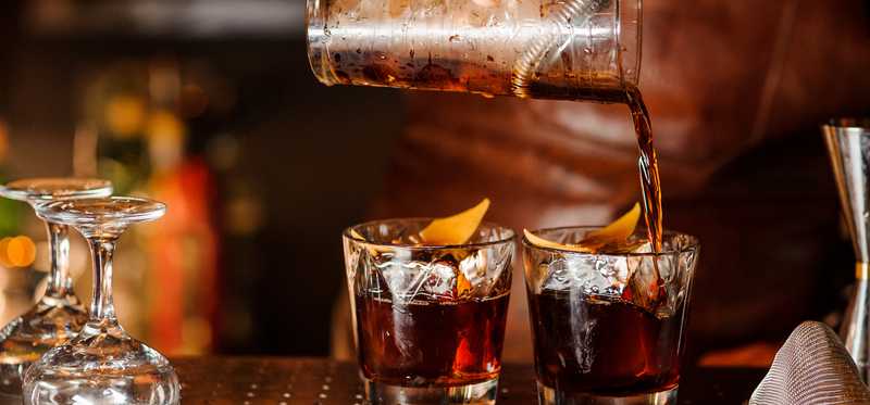 A bartended pours two drinks into glasses at a bar.