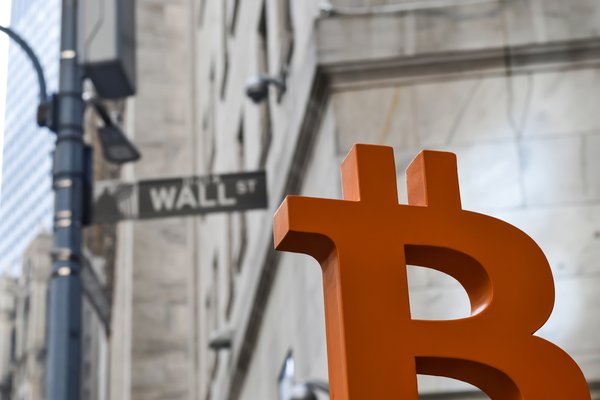 A large, red Bitcoin symbol stands next to a New York street sign marked Wall Street.