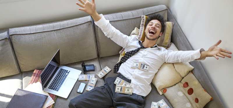 Businessman lying back on couch next to computer and shouting for joy while throwing cash in the air