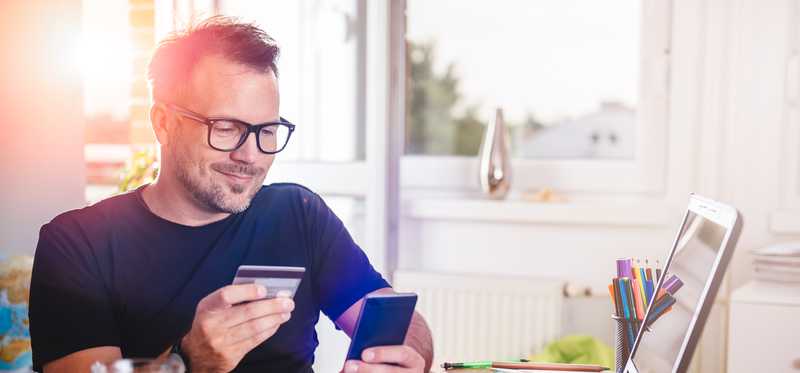 A man holds a credit card and a smartphone in front of a laptop.