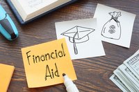 Financial aid written on a Post-It note lying on a desk next to a pile of hundred dollar bills and drawings of a cap and a bag of money.
