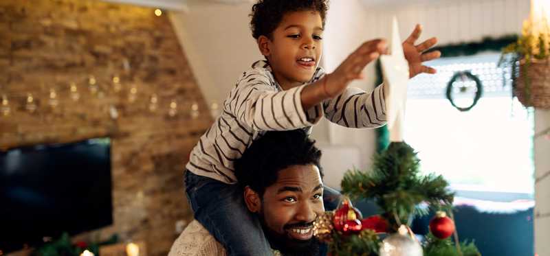 Child on adult's shoulders is putting star on Christmas tree.