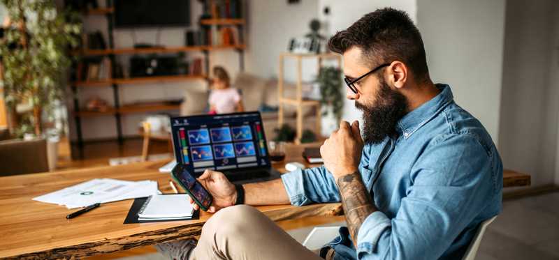Adult holding smartphone sits at desk with tablet showing multiple charts.