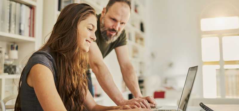 Person smiles while typing on laptop as another person stands by to help.