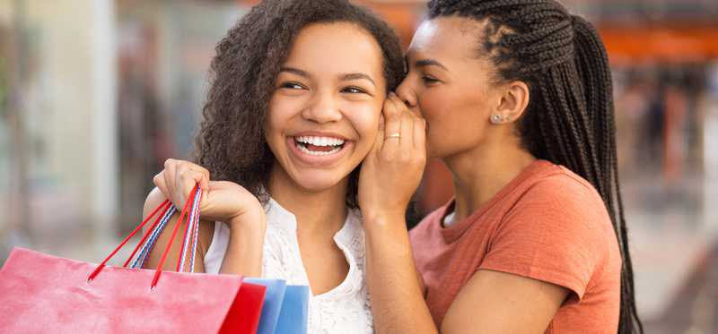 One smiling person whispering in the ear of another who is carrying shopping bags.