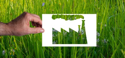 Green wash: A man's hand holds a blank sheet of paper with the silhouette of a factory cut out. Green grass background. Not everything "green" is ecological