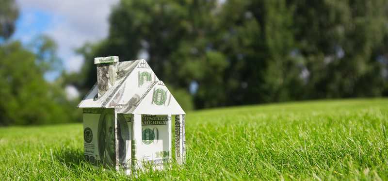 A house made of folded U.S. dollar bills sits on a grassy lawn.