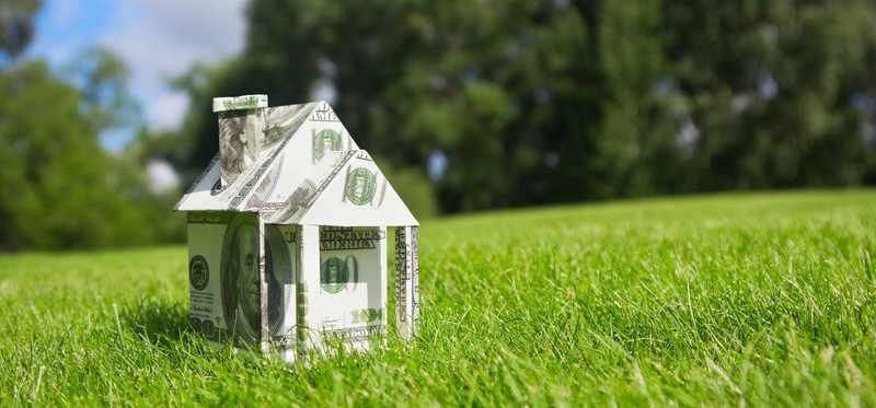 A house made of folded U.S. dollar bills sits on a grassy lawn.
