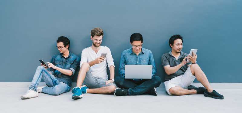 Four millennials sit against a wall all using electronic devices.
