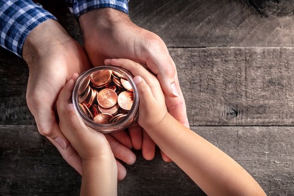 A child's hands grasp a jar of pennies and their hands rest in an adult's hands.