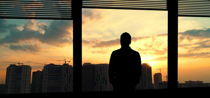 Person standing and looking out window at sunset over city.