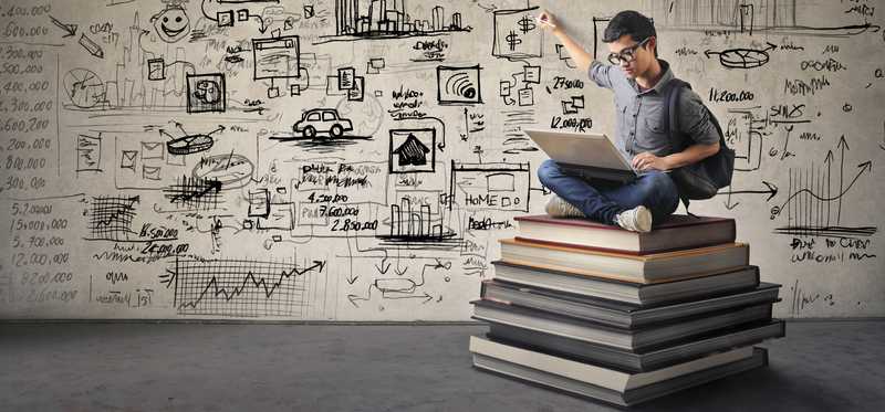 Man in glasses sitting on a stack of books pointing to a wall with writing all over it behind him.