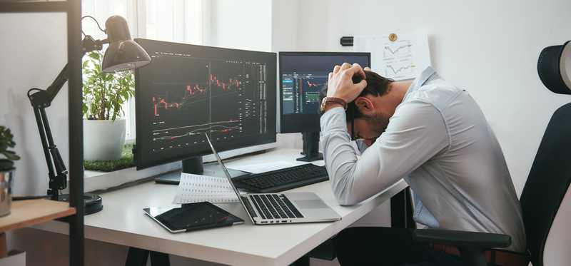 Frustrated investor holds his head in his hands in front of computer screens.