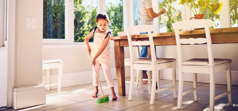 Child sweeps floor while adult cleans window in sunny room.