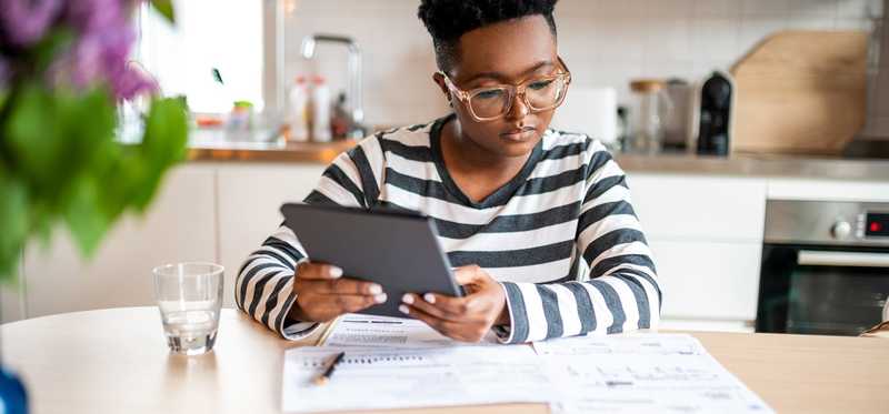 Person sitting at a kitchen table and looking at papers while holding a tablet.