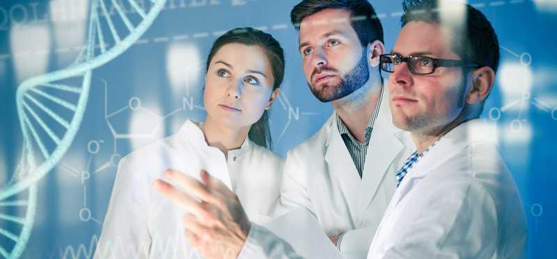 Researchers in white coats work together in front of a monitor displaying a double helix.
