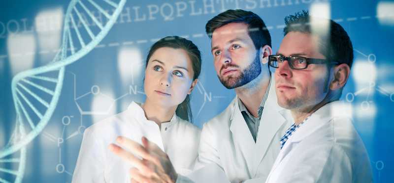 Researchers in white coats work together in front of a monitor displaying a double helix.