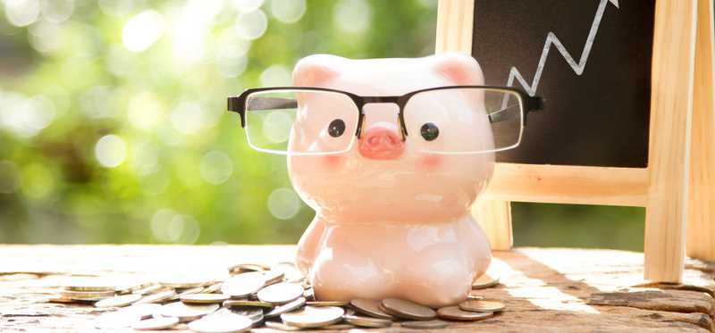 Small piggy bank wearing glasses sitting on a pile of coins with small chalkboard rising chart in the background.